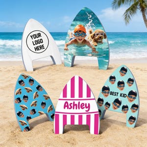 May include: Five surfboard-shaped foam kickboards standing on sand. One features a logo placeholder, one shows a child and dog swimming, one has a repeating pattern of faces, one is pink and white striped with the name Ashley, and one has faces with the text Best Kid.