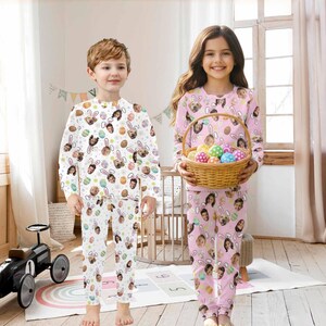 May include: Two children wearing matching Easter-themed pajamas. The pajamas feature a pattern of faces, Easter eggs, and bunny ears. One child wears white pajamas, the other wears pink pajamas and holds a basket of colorful Easter eggs.