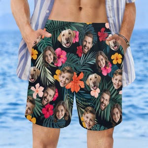 May include: Black swim trunks with a tropical floral and face pattern. The design includes faces, flowers, and palm leaves. The trunks have a drawstring waist and pockets.