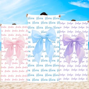 May include: Three personalized beach towels with large watercolor bows. One towel has a pink bow and the name "Amelia" repeated. Another has a blue bow and the name "Olivia". The third has a purple bow and the name "Evelyn".