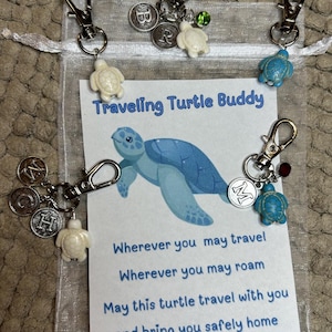 May include: A set of three turtle keychains in a white mesh bag. The keychains feature a white turtle charm with a silver letter charm and a blue turtle charm with a silver letter charm. The bag has a card with the text "Traveling Turtle Buddy" and a poem about traveling safely.