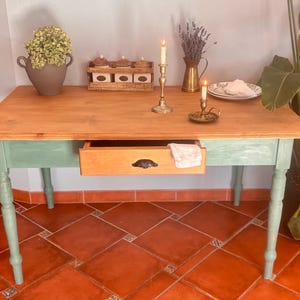 Puede incluir: Mesa de madera con tapa de madera natural y patas y faldón pintados de verde menta. La mesa tiene un solo cajón con un tirador negro. Elementos decorativos incluyen velas, flores y un plato.