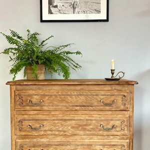 Puede incluir: Una cómoda de madera de tres cajones con tallas ornamentadas y tiradores plateados. La cómoda es de color marrón claro y tiene un acabado desgastado. Está sentada sobre un suelo de baldosas frente a una pared con una fotografía en blanco y negro enmarcada del Puente de la Torre en Londres.