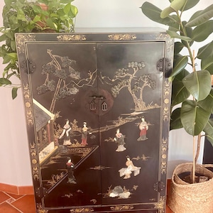 Könnte beinhalten: Ein schwarzer Lackschrank mit zwei Türen und goldfarbenen Metallbeschlägen. Die Frontplatten sind mit detaillierten, erhabenen Figuren in traditioneller Kleidung, Bäumen und architektonischen Elementen verziert, die von goldfarbenen Rändern umrahmt sind.