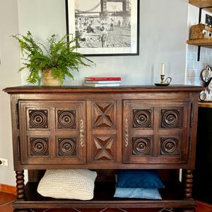 Puede incluir: Un mueble de madera oscura con diseños geométricos tallados en las puertas. Una fotografía en blanco y negro enmarcada cuelga encima. Una planta en maceta y libros están encima. Dos almohadas son visibles en el estante inferior.