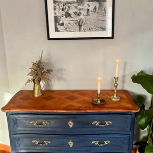 Könnte beinhalten: Eine marineblaue antike Kommode mit Holzplatte und Messingbeschlägen. Die Oberseite hat ein geometrisches Muster. Zwei Messingkerzenhalter und eine Vase mit getrockneten Blumen stehen oben. Ein schwarz-weiß gerahmtes Bild hängt darüber.
