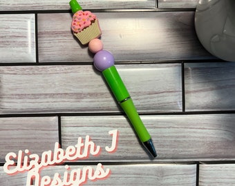 Op de afbeelding: Een groene pen met een roze cupcake-bedel en paarse en roze kralen. De pen ligt op een tegelvloer met houtlook. De tekst "Elizabeth J Designs" is zichtbaar in de linkerbenedenhoek van de afbeelding.