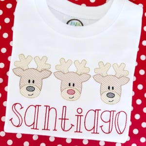 May include: White long-sleeve shirt with three embroidered reindeer faces and the name "Santiago" in red thread.
