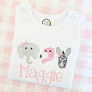 May include: White sleeveless onesie with pink, gray, and black embroidered animals. The animals are an elephant, a flamingo, and a zebra. The onesie has the name "Maggie" embroidered in pink below the animals.