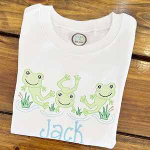 May include: White t-shirt featuring three embroidered green frogs in a pond scene with grass and water details. The name "Jack" is embroidered in light blue script below the frogs. A small label is visible near the neckline.