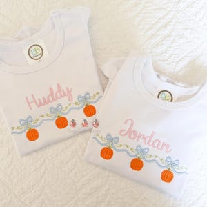 May include: Two white short-sleeved shirts featuring embroidered designs. One shirt displays the name Huddy in pink, and the other displays the name Jordan in pink. Both shirts feature a light blue ribbon garland with orange pumpkins and green vines.