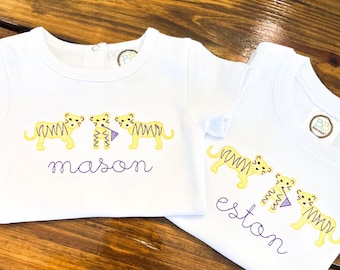 May include: Two white baby shirts with embroidered yellow tigers and purple names. One shirt says "mason" and the other says "eston". Each shirt has a small tag with the initials "BB" on it.