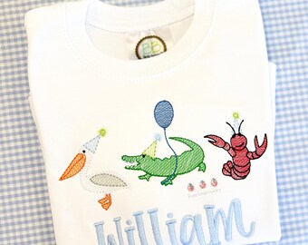 May include: White t-shirt with embroidered birthday-themed animals: a pelican, alligator, and lobster, each wearing a party hat. A blue balloon and the name "William" are also embroidered on the shirt. The shirt is folded on a blue and white checkered surface.