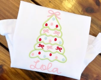 May include: White t-shirt with a green, pink, and red embroidered Christmas tree with the name "Lola" underneath.