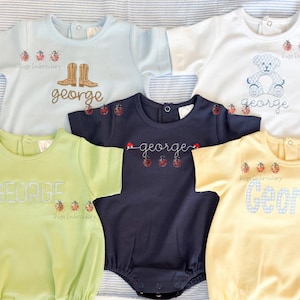 May include: Five short-sleeved rompers in light blue, white, navy, lime green, and yellow. Each features embroidered designs including ladybugs, cowboy boots, or a teddy bear, and the name George. The text Dugs Embroidery is visible on several.