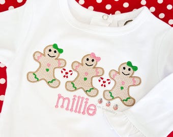 May include: White long-sleeved shirt with three embroidered gingerbread men, each with a different colored bow and candy cane. The name "millie" is embroidered in pink script below the gingerbread men. The shirt has a ruffled sleeve.