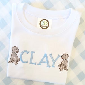 May include: White baby onesie with blue embroidered letters spelling "CLAY" and two brown dog illustrations on either side of the name. The onesie has a small tag with the text "BB" and "blink boutique" on it.