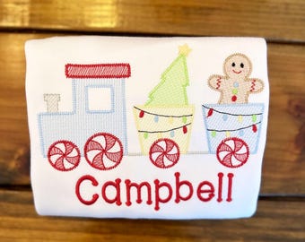 May include: A white onesie with a red, blue, and green embroidered design of a train pulling a Christmas tree and a gingerbread man. The train has candy cane wheels and the cars have lights. The onesie is personalized with the name "Campbell" in red embroidery.