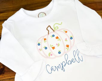 May include: White long-sleeved shirt featuring a pink embroidered pumpkin design. The pumpkin is decorated with blue, orange, and pink floral accents. The name "Campbell" is embroidered in blue script below the pumpkin.