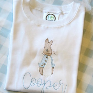 May include: White baby t-shirt with a blue embroidered Peter Rabbit design and the name "Cooper" embroidered below. The shirt has a small label with the text "BB Baby Boutique" on the top.