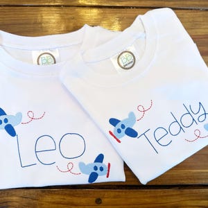 May include: Two white t-shirts with blue airplane designs and embroidered names "Leo" and "Teddy".