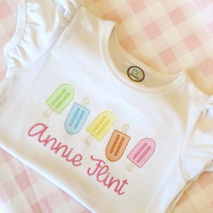 May include: White baby onesie with a row of five embroidered popsicles in rainbow colors. The onesie has the name "Annie Flint" embroidered below the popsicles.