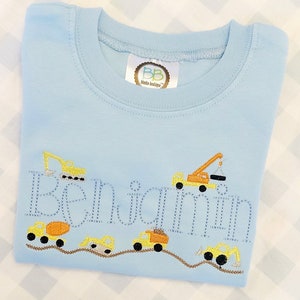 May include: Light blue long-sleeve shirt with embroidered construction vehicles and the name "Benjamin" in a dotted font. The shirt features a crew neck and a small tag at the neckline.