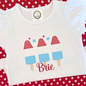 May include: White t-shirt with red, white, and blue popsicle graphic and the name "Brie" embroidered on the front.