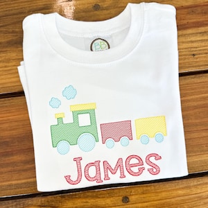 May include: White t-shirt featuring a colorful train design with the name "James" embroidered in red. The train is green, red, and yellow with blue accents. The shirt is folded on a wooden surface.