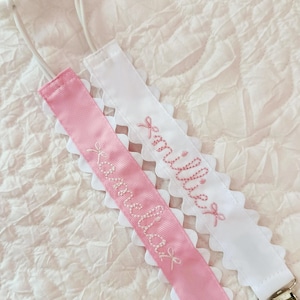 May include: Pink and white baby pacifier clips with embroidered names. The pink clip says "Amelia" and the white clip says "Millie". Both clips have a scalloped lace edge and a metal clip.