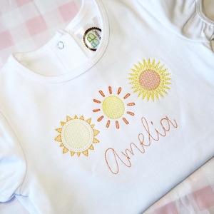 May include: White baby onesie with three embroidered suns in yellow, orange, and red. The name Amelia is embroidered in orange cursive below the suns.