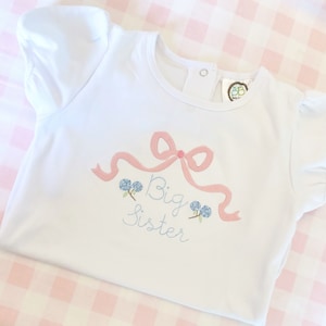 May include: White short-sleeved shirt with a pink bow and blue flowers embroidered on the front. The text "Big Sister" is embroidered below the bow.