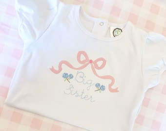 May include: White short-sleeved shirt with a pink bow and blue flowers embroidered on the front. The text "Big Sister" is embroidered below the bow.