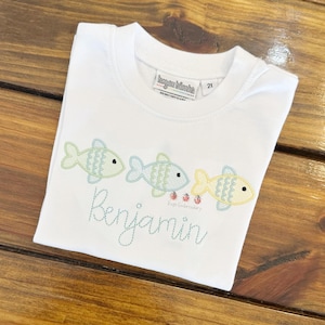 May include: White t-shirt with a crew neck featuring embroidered fish in pastel colors. The name "Benjamin" is stitched in cursive below the fish. The shirt has a tag that says "hagan blanks" and "2T".