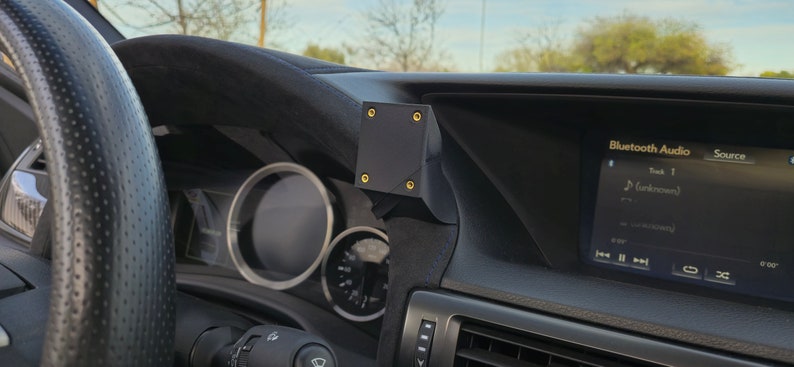 May include: A black car dashboard with a black phone mount attached to the left side. The mount has four gold screws. The car's dashboard screen displays "Bluetooth Audio" and "Source" with a song title and artist.