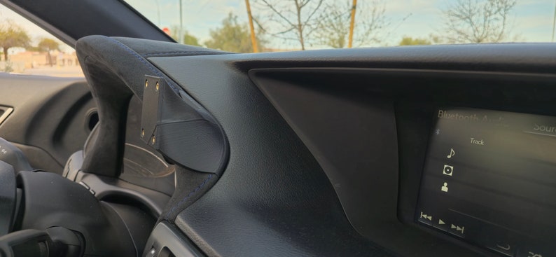May include: A black car interior with a phone mount attached to the dashboard. The car's infotainment screen displays "Bluetooth Audio" and "Track" with music note icon.