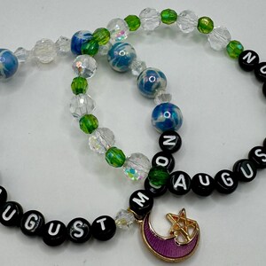 May include: Two beaded bracelets. One bracelet has blue and white marbled beads, clear beads, and black letter beads spelling "AUGUST". The other bracelet has green and clear beads, blue and white marbled beads, and black letter beads spelling "MOON". Both have moon charms.