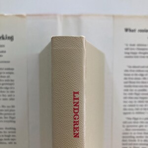 May include: A beige book spine with a textured surface featuring the name LINDGREN printed in red capital letters. The book is positioned vertically against a blurred background showing parts of a dust jacket with printed text on both sides.