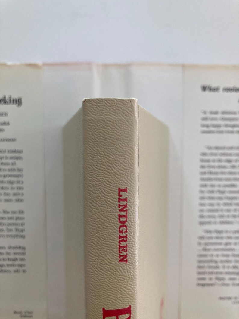 May include: A beige book spine with a textured surface featuring the name LINDGREN printed in red capital letters. The book is positioned vertically against a blurred background showing parts of a dust jacket with printed text on both sides.