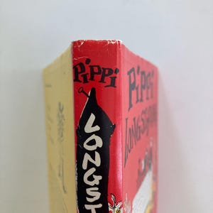 May include: A hardcover book titled Pippi Longstocking with a red spine featuring black text and a black silhouette illustration. The book has a yellow back cover and shows signs of wear along the top edge of the spine with white paper visible.