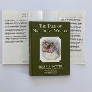May include: A hardcover book titled The Tale of Mrs. Tiggy-Winkle by Beatrix Potter with a dark green cover featuring an illustration of a hedgehog in a pink dress and white apron, displayed with its open dust jacket showing printed text.