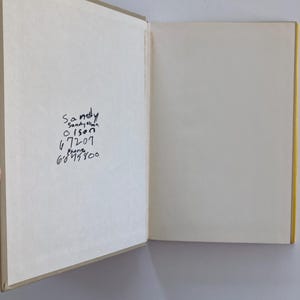 May include: An open hardcover book with beige endpapers. The left side features handwritten black ink text that reads: Sandy, Sandy Olson, Olson, 67207, Phone, 6845800. The right side is a blank, off-white page. The book has a yellow spine visible.