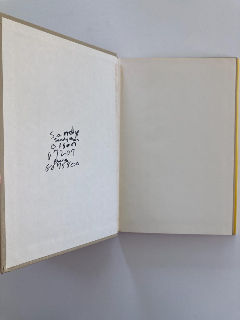 May include: An open hardcover book with beige endpapers. The left side features handwritten black ink text that reads: Sandy, Sandy Olson, Olson, 67207, Phone, 6845800. The right side is a blank, off-white page. The book has a yellow spine visible.