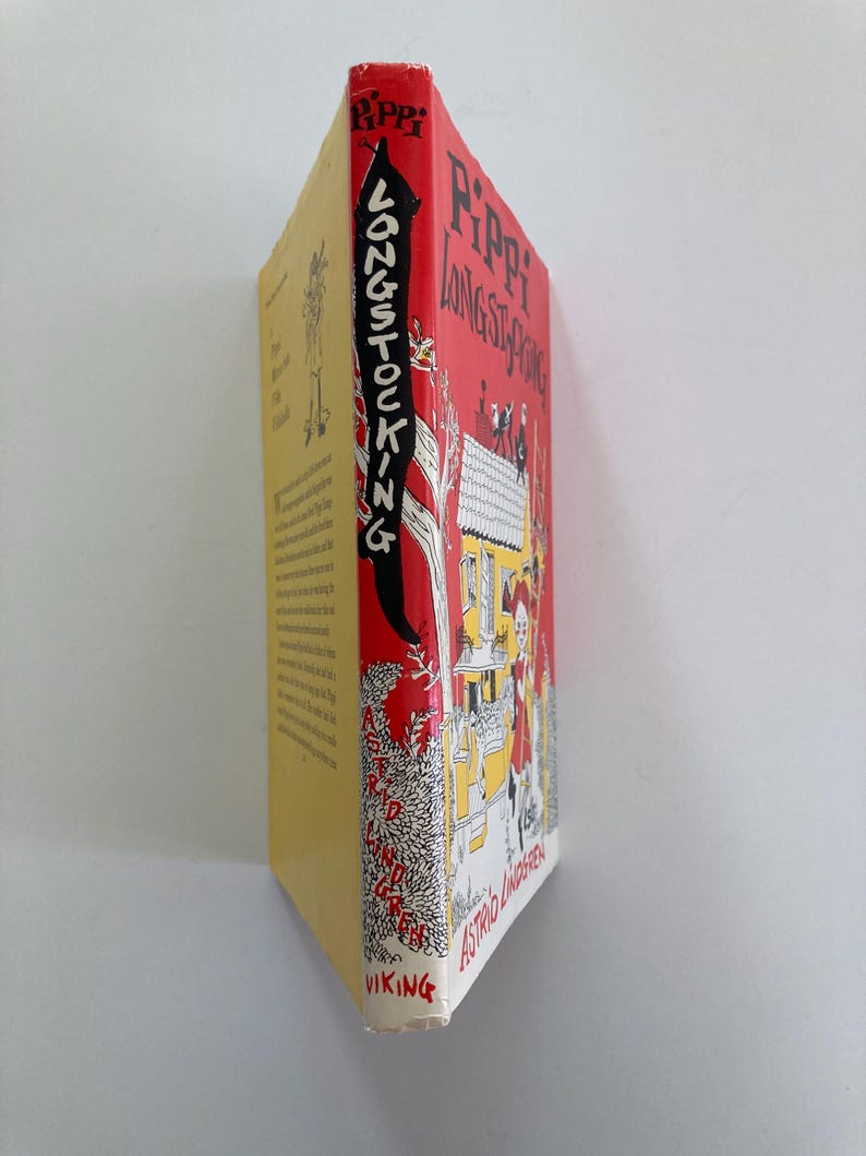 May include: A hardcover book titled Pippi Longstocking by Astrid Lindgren. The spine is red with black text and a white illustration of a tree. The front cover features a yellow house, a character with red hair, and black text on a red background.