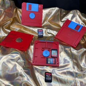 Peut inclure: Une disquette rouge et bleue avec une étiquette bleue et une cartouche de jeu rouge et bleue. La disquette est sur une surface en tissu dorée. La cartouche de jeu est sur un support de cartouche de jeu rouge et bleu.