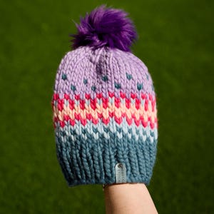 May include: A hand-knitted beanie in shades of purple, pink, and teal, topped with a fluffy purple pom-pom. The hat features a chevron pattern and a small tag with text. A cozy winter accessory.