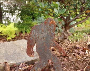 May include: A rusty metal silhouette of Bigfoot walking through a garden with brown mulch and green foliage.