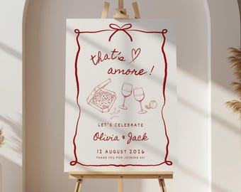 May include: A white sign on a wooden easel with a red border and text. The sign says "that's amore!" and "Let's Celebrate Olivia & Jack 12 August 2026". The sign also features illustrations of pizza, wine glasses, and a tomato.