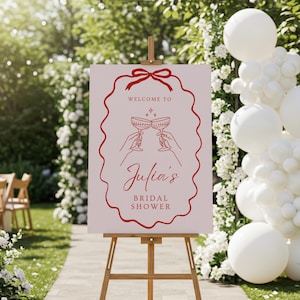 Może przedstawiać: Jasnoróżowy znak na drewnianym sztaludze z napisem "Welcome to Julia's Bridal Shower" w czerwonym skrypcie. Znak posiada czerwoną ozdobną obwódkę i ilustrację dwóch kieliszków do szampana.