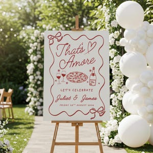 Może przedstawiać: Biały znak na drewnianym sztaludze z napisem "That's Amore" w czerwonym skrypcie, wraz z ilustracjami kieliszków do wina, pizzy i butelki. Na znaku widnieje napis "Let's Celebrate Juliet & James Friday 20th August 2026."